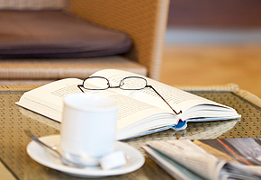 Auf einem Tisch steht eine Tasse Kaffee und danaben liegt ein Buch, eine Zeitung und eine Brille.
