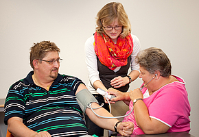 Eine Therapeutin erklärt einer Patientin, wie sie einem anderen Patienten den Blutdruck messen kann.